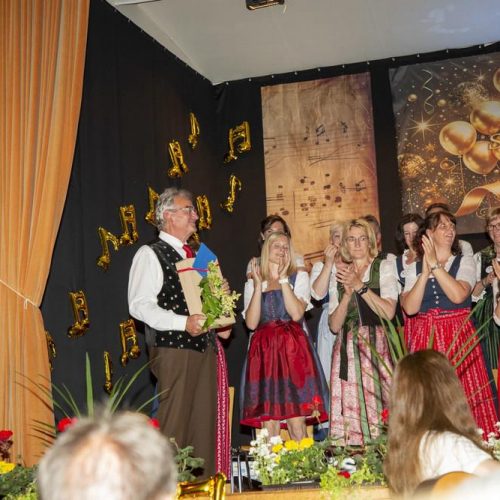 Der Chorleiter Richard Pertl hält einen Blumenstrauß, während die Sängerinnen auf der Bühne applaudieren.